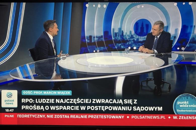 Dwie osoby siedzą przy stole w studiu telewizyjnym i rozmawiają. 