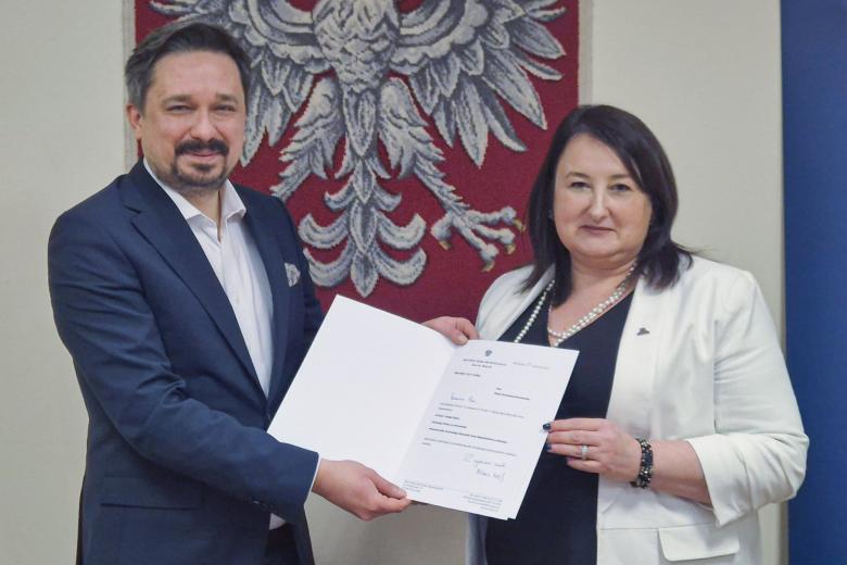Marcin Wiącek i Beata Tarnawska-Domalewska pozują do zdjęcia, uśmiechając się i pokazując w stronę aparatu otwartą teczkę z dokumentem powołania