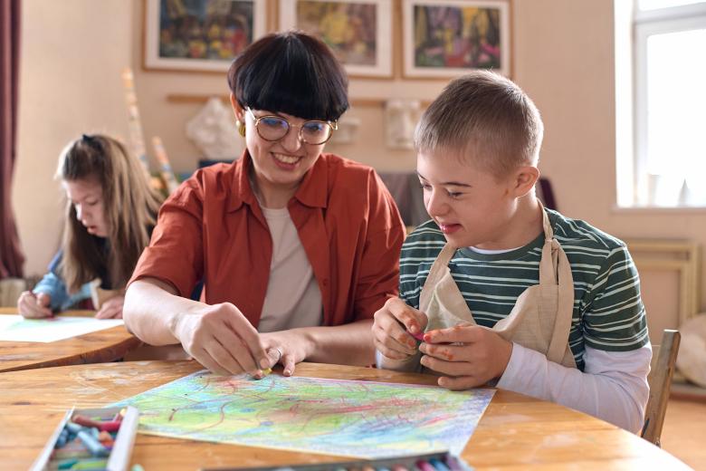 Przy stole siedzą uśmiechnięci opiekunka i dziecko z zespołem Downa, wspólnie malując pastelami na kartce papieru