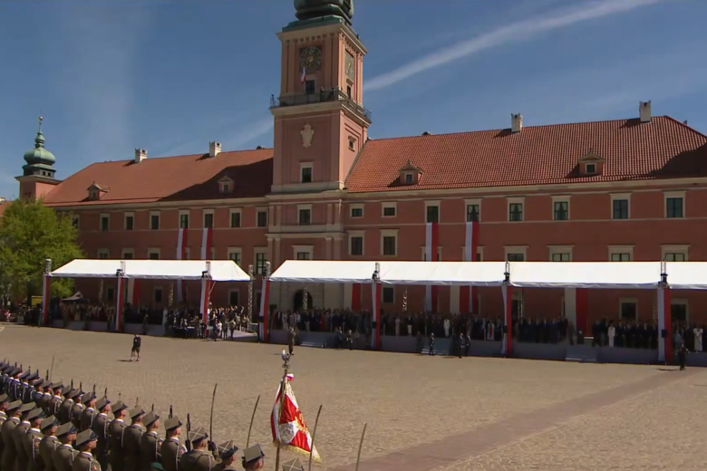 Plac przed Zamkiem Królewskim w Warszawie. Fasada zamku przyozdobiona jest biało-czerwonymi flagami. Pod fasadą stoi długi namiot z trybunami dla uczestników uroczystości. Po przeciwległej stronie placu w dwuszeregu stoją na baczność żołnierze.