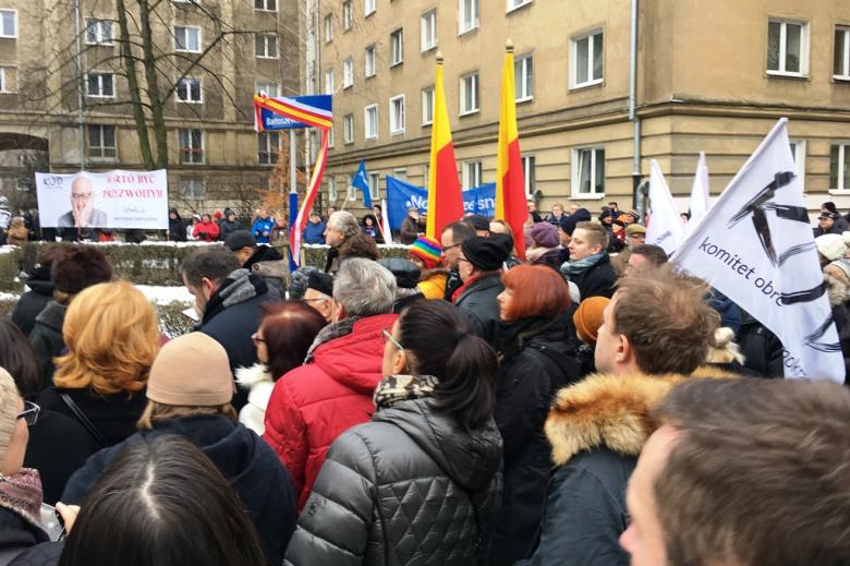 Zdjęcie: tłum ludzi przed budynkiem z lat 50., flagi Warszawy i banner z wizerunkiem Władysława Bartoszewskiego