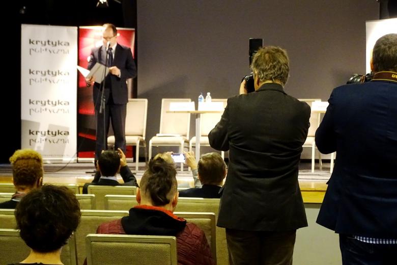 zdjęcie: na scenie stoi mężczyzna w garniturze, przed nim siedzi kilka osób, po prawej stronie stoi dwóch fotografów