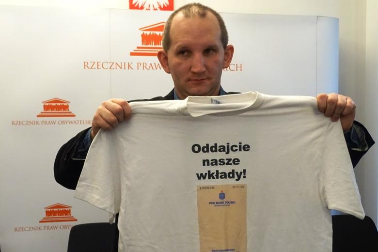 zdjęcie: mężczyzna trzyma w rękach rozłożoną koszuklkę z napisem: Oddajcie nasze wkłady