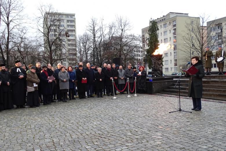 zdjęcie: przed pomnikiem przy mikrofonie stoi kobieta, obok stoi kilkadziesiąt osób