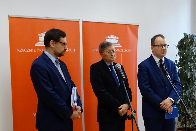 zdjęcie: trzech mężczyzn w garniturach stoi na tle pomarańczowych banerów