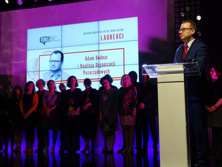 na zdjęciu dr Adam Bodnar stoi przy pulpicie, za nim w tle stoją przedstawiciele organizacji pozarządowych