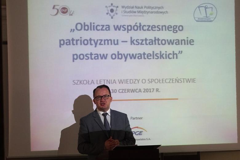 zdjęcie: mężczyzna w jasnym garniturze i okularach stoi przy mównicy