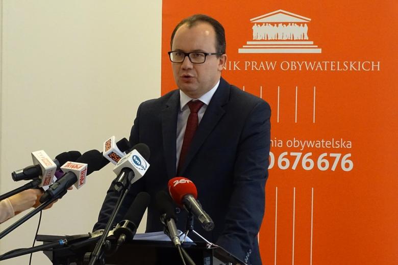 zdjęcie: mężczyzna w garniturze stoi przy mównicy, przed nim stoi kilka mikrofonów