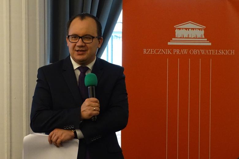 zdjęcie: mężczyzna w garniturze stoi na tle biało-pomarańczowego baneru