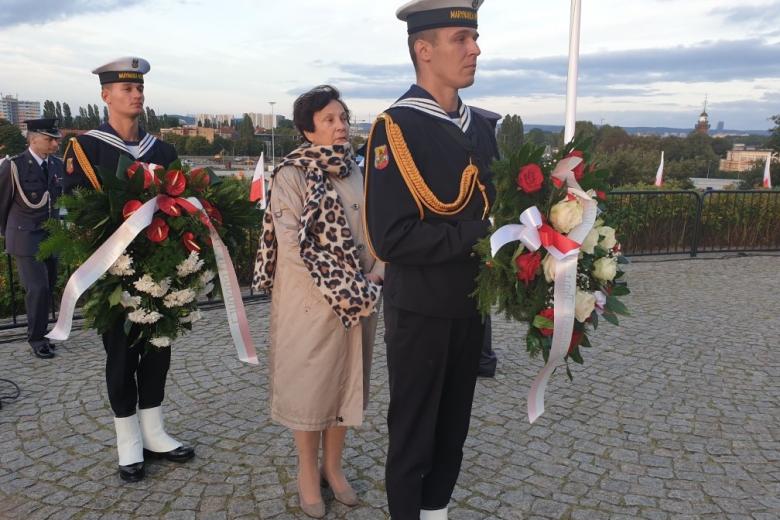 mężczyzna w mundurze trzyma wieniec, za nim kobieta w płaszczu
