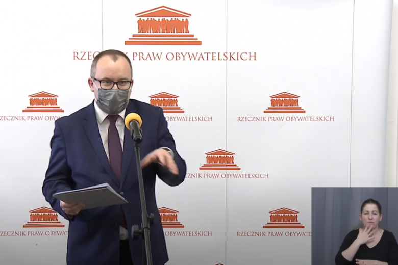 Mężczyzna w garniturze w maseczce przed mikrofonem. W rogu sylwetka tłumaczki PJM