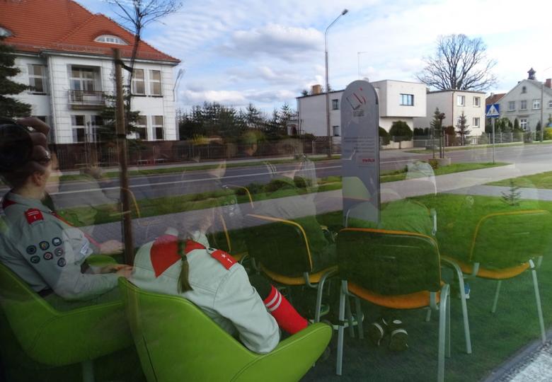 Widok na salę biblioteczną z ulicy. Widać ludzi, a w szybie odbija się ulica