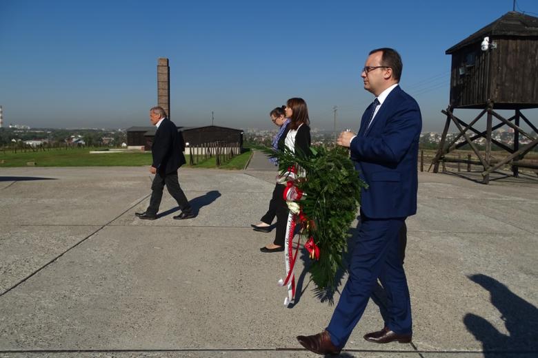 Zdjęcie: grupa ludzi z wieńcem w barwach narodowych, w tle barak obozowy i krematorium