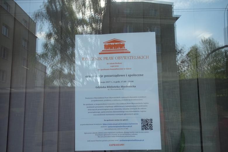 Plakat informacyjny na szybie, odbijają się w niej budynki z lat 60. i 50. XX wieku