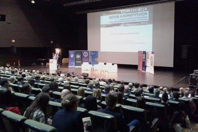 zdjecie: kilkadziesiąt osób siedzi na sali, przed nimi scena, na której stoi mężczyzna