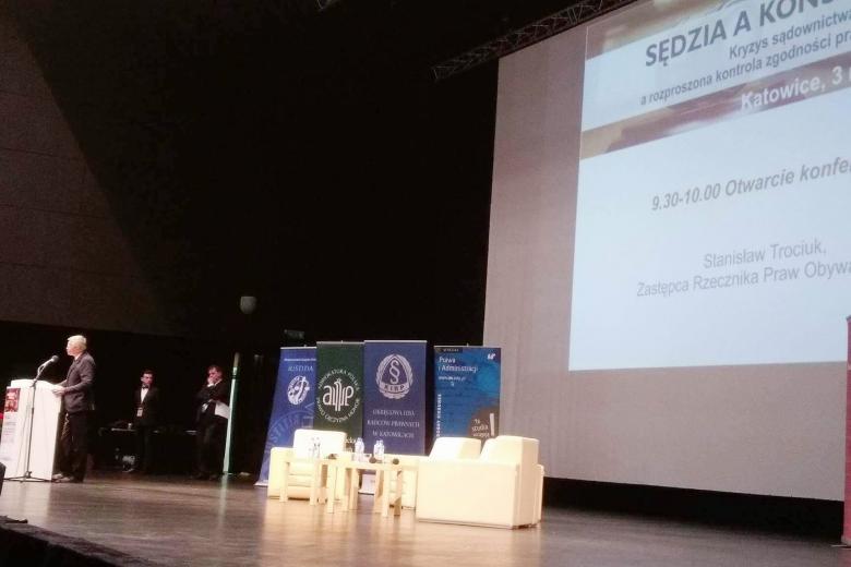 zdjęcie: na scecnie po lewej stronie stoi mężczyzna przy mównicy, za nim duży ekran