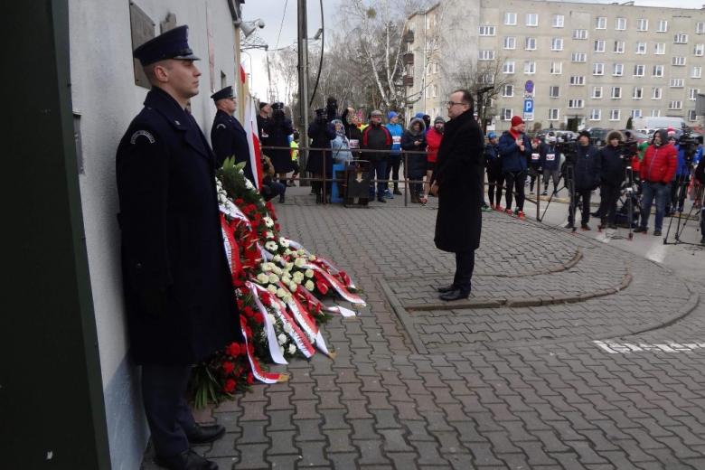 zdjęcie: mężczyzna stoi przed tablicą pod którą leżą biało-czerwone kwiaty