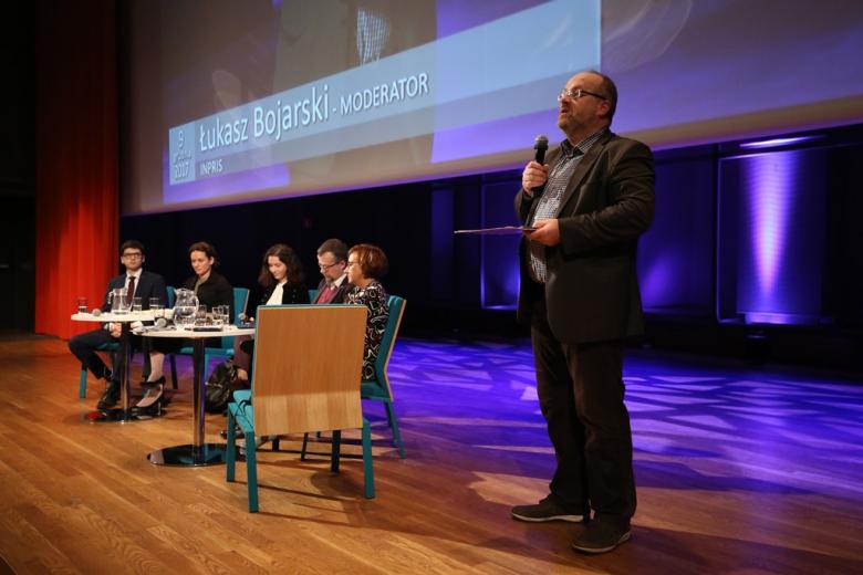 Mężczyzna stoi na scenie. Obok paneliści