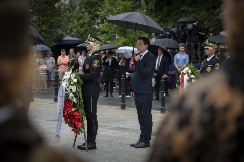 kobieta w mundurze trzyma wieniec, za nią mężczyzna z parasolem