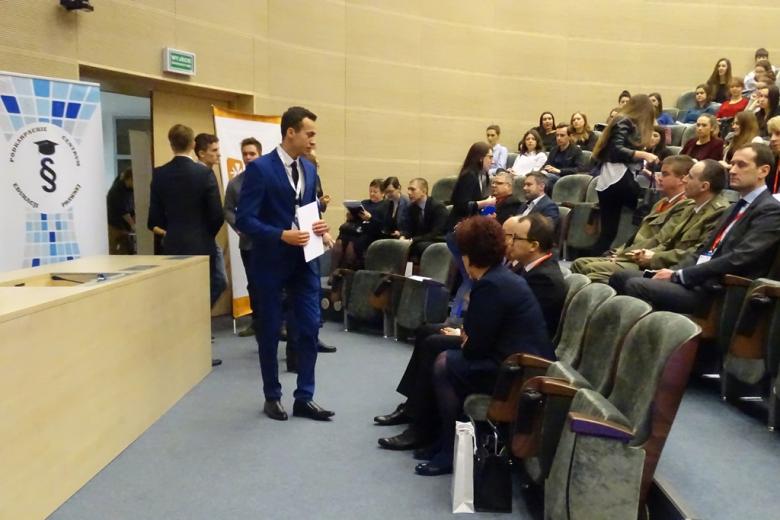 Zdjęcie: młody mężczyzna stoi i rozmawia z gośćmi konferencji siedzącymi w pierwszym rzędzie