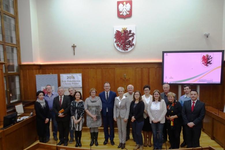 Zdjęcie grupowe na sali sejmiku. Godło państwa, herb województwa i krzyż na ścianie