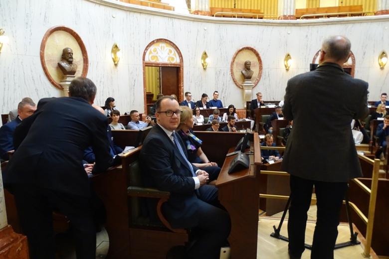 Sala posiedzeń. Mężczyzna patrzy w obiektyw siedząc w ławie dla gości