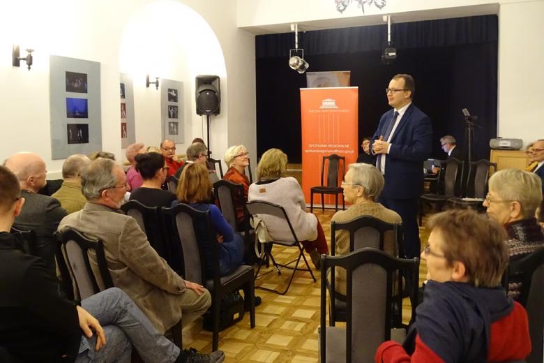 Zdjęcie: ludzie siedzą w zabytkowej sali, mężczyzna stoi