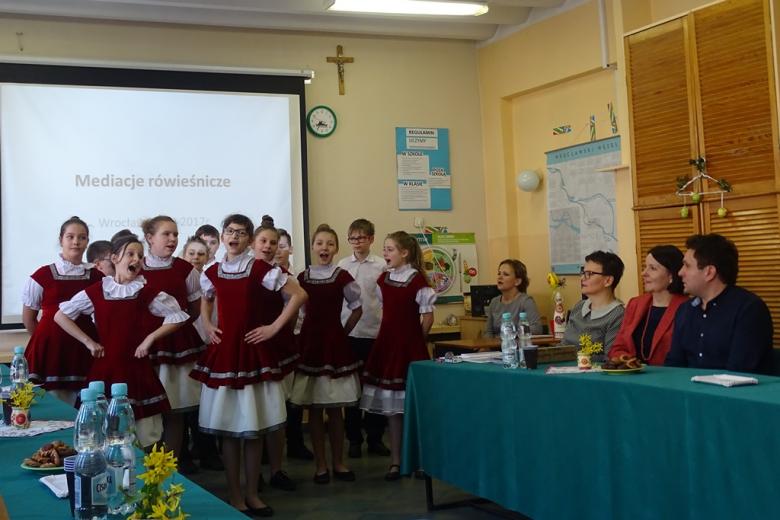 Dzieci w amarantowo-białych strojach śpiewają, goście siedzą przy zielonym stole 