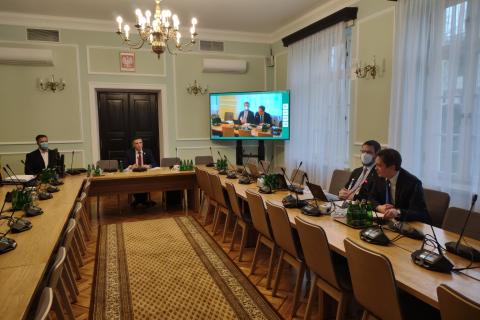 Kilka osób siedzi przy długich stołach konferencyjnych, w tle ekran z uczestnikami online