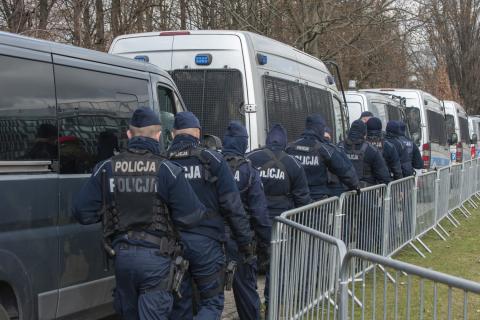 Kilkunastu policjantów idzie wzdłuż barierek oddzielających trawnik od ulicy, na której stoją zaparkowane radiowozy policyjne