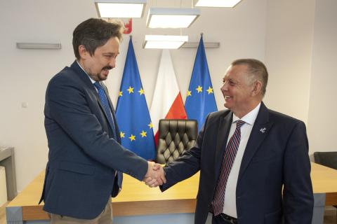 RPO Marcin Wiącek i prezes NIK Marian Banaś pozują do zdjęcia na tle flag Polski i UE ściskając dłonie na powitanie