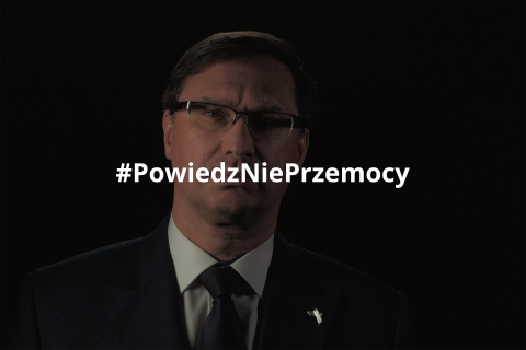 Robert Damski stojący na czarnym tle i spoglądający w obiektyw. Na obraz nałożony jest hashtag "Powiedz Nie Przemocy"