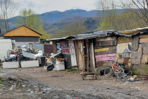 Podniszczone, drewniane szopy z desek, przykryte folią, przed szopami sterta śmieci, stary rower, błotnista droga