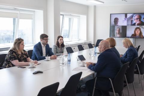 Sześć osób siedzi po dwóch stronach długiego, prostokątnego stołu, na długim boku duży ekran z uczestnikami spotkania łączącymi się za pomocą programu do komunikowania się na odległość