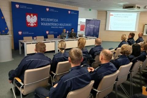 Grupa kilkudziesięciu policjantów w mundurach słucha dwóch osób siedzących przy stole w dużej sali konferencyjnej