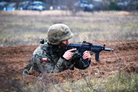 Żołnierz w hełmie i pełnym umundurowaniu leży na ziemi i celuje z karabinku automatycznego