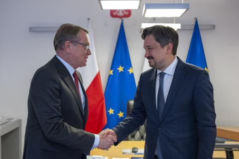 Dwie osoby witają się ściskając dłonie na tle flag Polski i UE