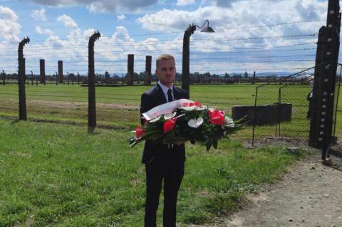 Mężczyzna w garniturze trzyma biało-czerwony wieniec na tle ogrodzenia obozowego