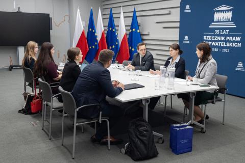 Siedem osób siedzi po dwóch stronach dużego prostokątnego stołu w sali konferencyjnej