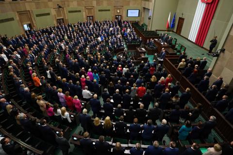 Duża sala posiedzeń z kilkuset osobami stojącymi w kilkunastu rzędach ław ułożonych w półokręgu