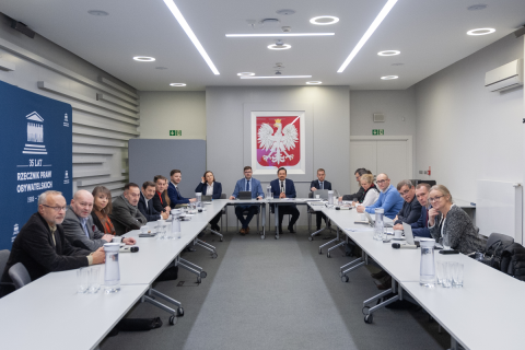 Kilkanaście osób siedzi przy stołach ustawionych w kształt podkowy na sali konferencyjnej, w tle godło Polski