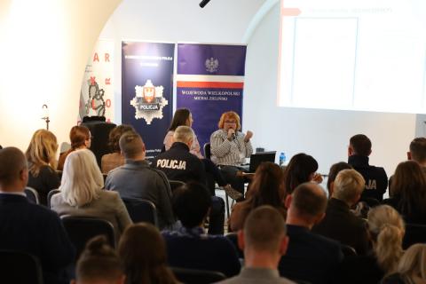 Kilkadziesiąt osób słucha wystąpienia dwóch kobiet siedzących na tle plakatów z napisem "Policja"