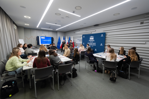 Kilkadziesiąt osób siedzących w grupach przy stołach w sali konferencyjnej słucha prowadzącego spotkanie