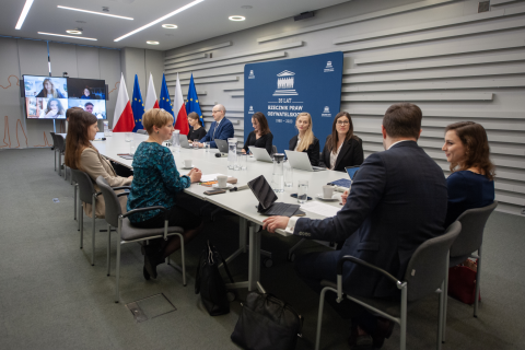 Kilkanaście osób siedzi przy stole konferencyjnym i rozmawia, w tle flagi Polski i UE