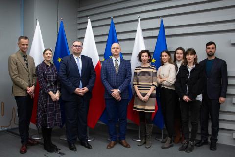 Grupa osób pozuje na stojąco do wspólnego zdjęcia na tle flag Polski i UE