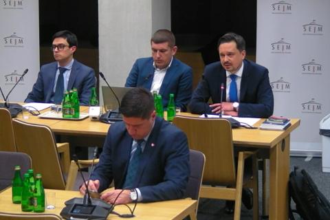 Marcin Wiącek, Wojciech Brzozowski i Michał Szwast siedzą przy stole w sali komisji sejmowej. Marcin Wiącek wypowiada się do mikrofonu.