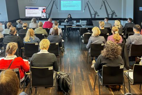 Kilkadziesiąt osób siedzi w sali szkoleniowej. W szczycie sali za stołem siedzi prelegentka prowadząca szkolenie, za której plecami wyświetla się prezentacja multimedialna