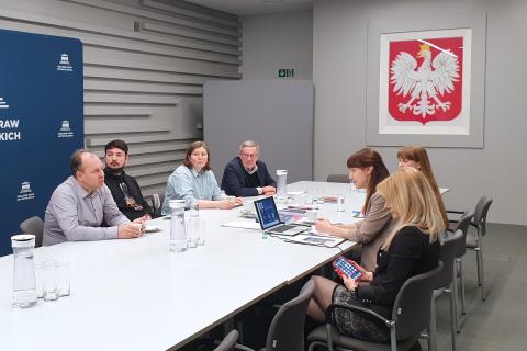 Siedem osób siedzi przy dużym stole w sali konferencyjnej i rozmawia