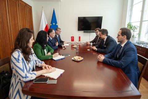 Siedem osób siedzi po obu stronach stołu i rozmawia. W tle flagi Polski i UE