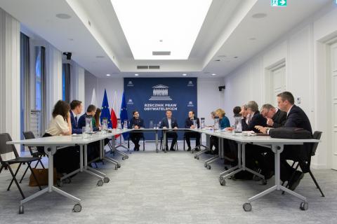 Dwanaście osób siedzi za stołami ustawionymi w podkowę na dużej sali konferencyjnej, w tle granatowa ścianka z białym napisem "Rzecznik Praw Obywatelskich"